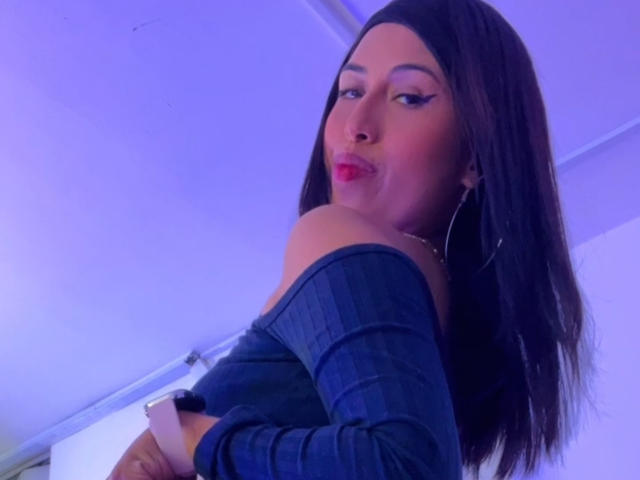 MyaaTyler with long brown hair, off-shoulder blue top, red lipstick, and hoop earrings, looking back with a slight pout, wearing a black wristband.