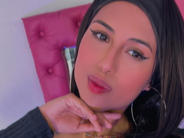 MyaaTyler with dark hair, pink eyeliner, and pink lips, holding her neck, wearing a gray top and hoop earrings, against a pink tufted background.