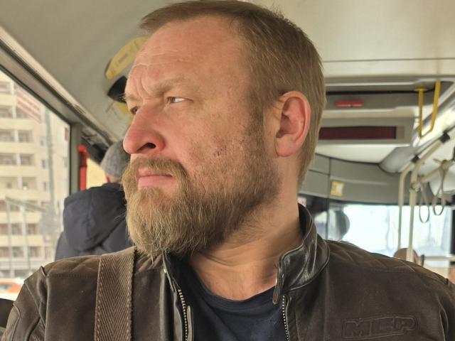 Grandor has a balding head with a scruffy beard, wearing a brown leather jacket, looking to the side with a serious expression inside a bus.