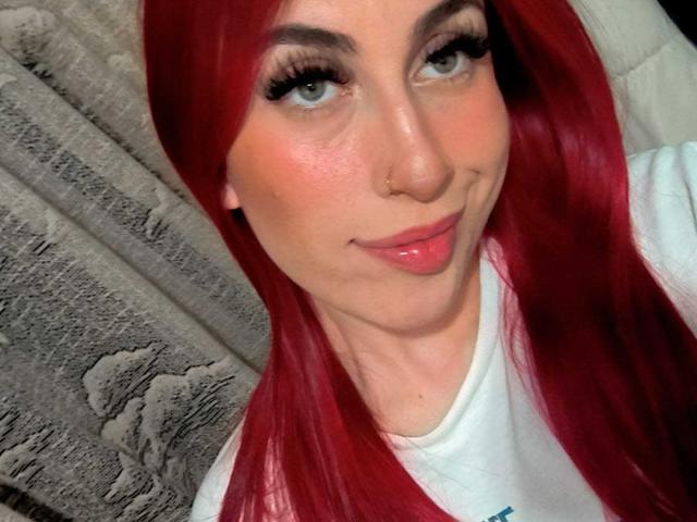 SammyViolet with red hair, blue eyes, and glossy pink lips, wearing a white shirt, nose ring, and black ear gauges, smiling slightly. Her ass is not visible.