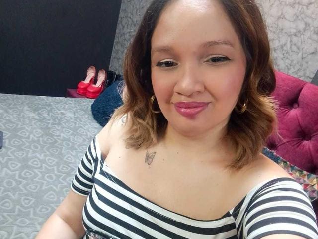 BiancaMorettii with brown wavy hair, wearing a black and white striped top, showing her tits, small tattoo above, pink lipstick, gold earrings, and a slight smile.