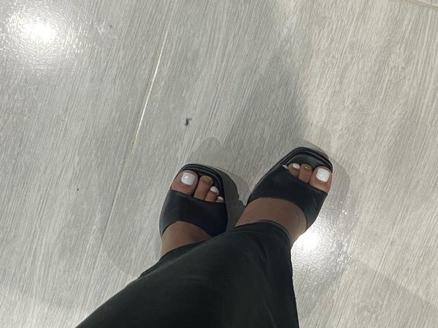 LucyLars' feet with black open-toe sandals, white-painted toenails, wearing black pants, standing on a shiny gray wooden floor.