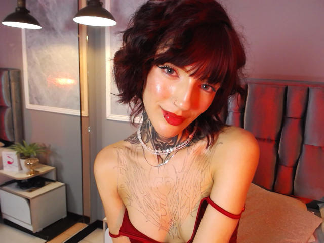 Tifany Diaz, com cabelo preto, tatuagens nos braços e na barriga, usando uma cropped preta que mostra seus pequenos seios e uma sunga preta, sentada em uma cama..