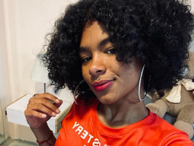 LucyLars, uma mulher de pele escura com longos cabelos pretos presos em um rabo de cavalo, vestindo lingerie vermelha de renda, um colar de pérolas e grandes brincos de argola, tem batom vermelho e uma tatuagem no peito..