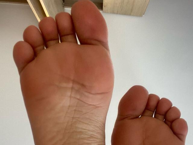 Las plantas de los pies de JazminSweet69 son el foco principal, mostrando piel arrugada y dedos ligeramente rojos, con su pie derecho más cerca y su pie izquierdo más abajo, bajo un techo de madera..