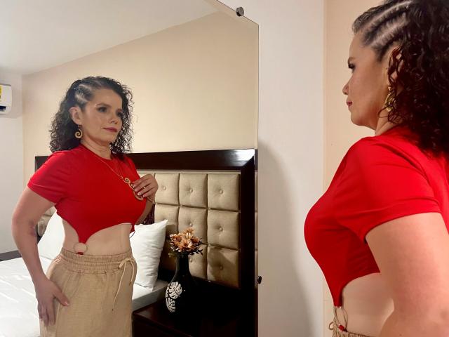 MadiFoxy with curly hair, red crop top showing her tits, beige pants, standing in front of mirror, looking at reflection, wearing gold earrings, in bedroom.