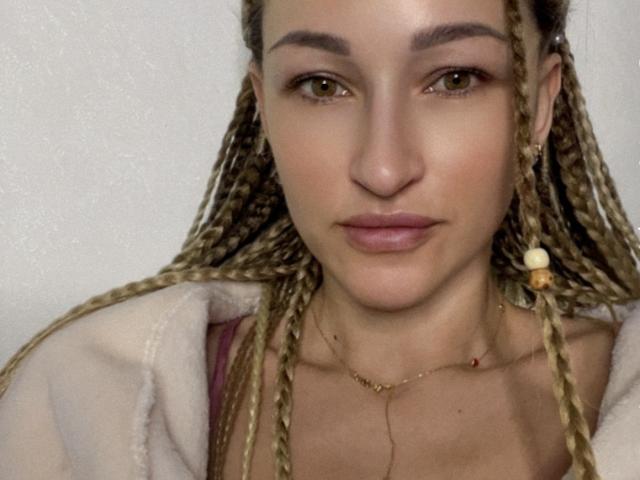 Lilitraya with long, blonde braids, green eyes, and light makeup, wearing a gold necklace and pink top, has a serious expression and a soft, white garment.
