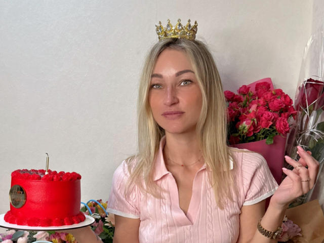 Lilitraya with blonde hair, wearing a pink striped shirt, small crown, holding red candlelit cake, pink roses, and gold bracelet, smiling. Her tits are small, face is pretty.