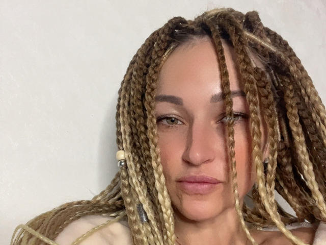 Lilitraya has light brown, braided, blonde-tipped hair, and a serious expression, with subtle makeup, and a slight smile, wearing a beige top, against a plain white background.