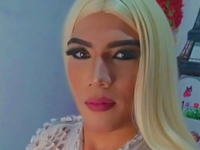 RoxanaLatinaTrans has platinum blonde hair, thick black eyebrows, dramatic eye makeup, and pink lipstick. She's wearing a white lace top with visible boobs.