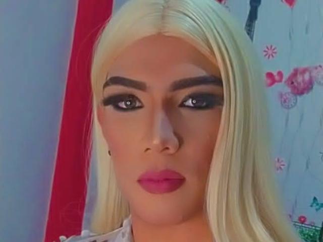 RoxanaLatinaTrans has blonde hair, thick eyebrows, dark eye makeup, and pink lips. She's looking directly at the camera with a serious expression. Her tits are not visible.