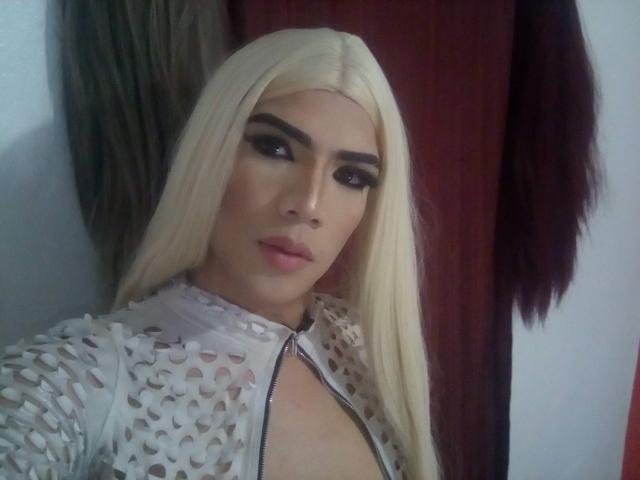 RoxanaLatinaTrans with long blonde hair, dark eyebrows, and heavy makeup, wearing a white, patterned, zipper-front top, exposing cleavage. Her lips are slightly parted, showing a subtle smile.