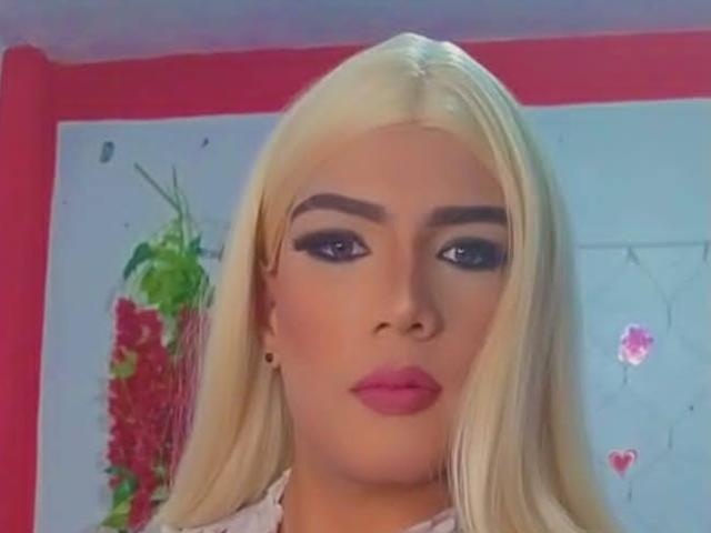 RoxanaLatinaTrans with blonde hair, full makeup, pink lipstick, and thick eyebrows, looking straight at the camera. Her tits are partially visible under a white top.
