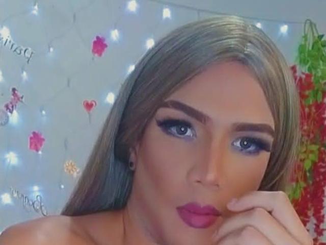 RoxanaLatinaTrans with long blonde hair, blue eyes, heavy eyeliner, purple lipstick, and black ear gauges, looking at the camera with her hand near her face. Boobs not visible.
