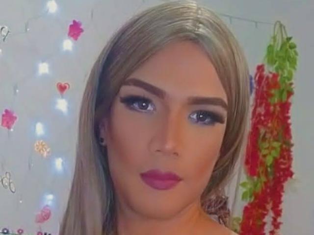 RoxanaLatinaTrans has blonde hair, purple lipstick, and thick eyebrows. Her blue eyes are slightly upturned. She's wearing black eyeliner and small black earrings.
