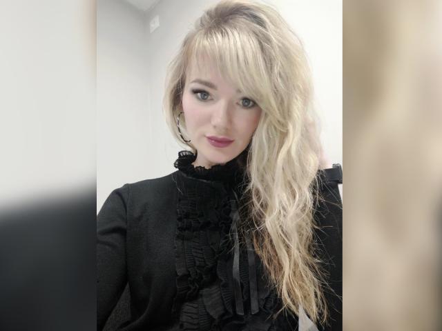 BrightBliss with long, wavy blonde hair, wearing a black ruffled top, has pink lipstick and subtle eye makeup, looking directly at the camera with a slight smile.