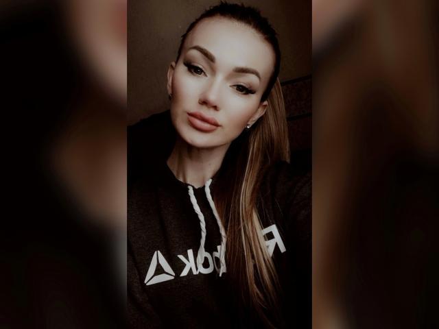 BrightBliss with long brown ponytail, wearing black hoodie, has pouty pink lips, subtle eyeliner, and diamond earrings, looking slightly to the side, with blurred brown background.