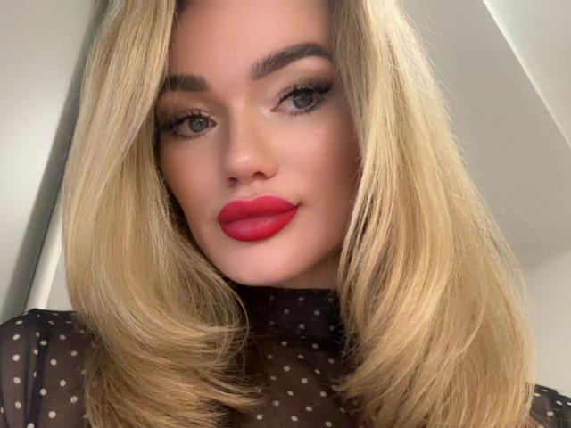 BrightBliss has blonde hair, red lipstick, and a sheer black polka dot top, with full eyebrows and long eyelashes, looking at the camera with a slight smile.