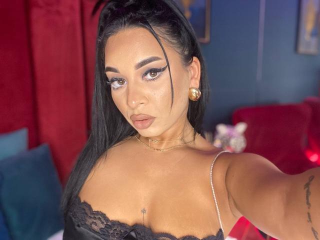 MadyLux mit schwarzer Spitzenbluse, die ihre Brüste zeigt, mit langen schwarzen Haaren, dicken Augenbrauen, goldenen Ohrringen und einem ernsten Gesicht, die ein Selfie in einem farbenfrohen Raum macht..