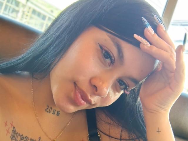 PerlaJones69 with black hair, blue eyes, and a tattoo on her neck. She's smiling, showing off her pink lips, and touching her head with long, patterned nails. Boobs visible.