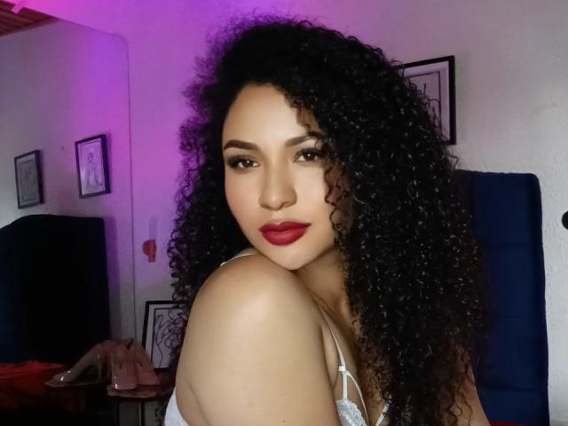 CarlaMuille with curly black hair, red lipstick, and brown eyes, looking over her shoulder, wearing a white lace top, showing her bare shoulder and upper chest.