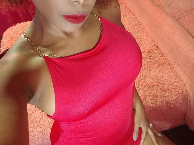 AfroditaKane vestindo um vestido vermelho justo com alças de corrente douradas, mostrando seus pequenos seios e traseiro curvilíneo, cama rosa e fofa ao fundo, batom vermelho, cabelo cacheado..
