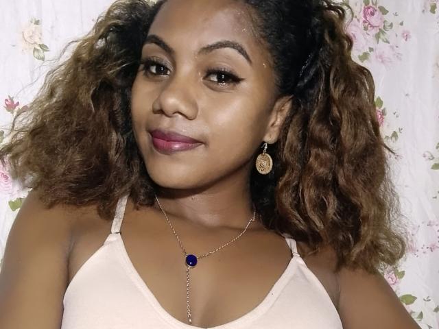 KennyReah with curly brown hair, brown skin, and pink lipstick, wearing a pink bra and blue pendant necklace, smiling slightly. Boobs are visible.