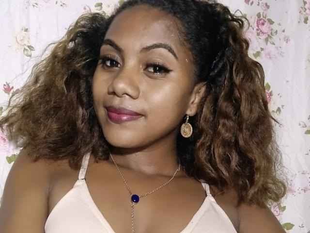 KennyReah with curly brown hair, dark eyebrows, pink lipstick, gold earrings, and a pink bra, wearing a silver necklace with a purple gem. KennyReah has a cute smile.
