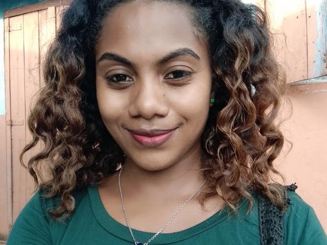 KennyReah has curly brown hair, medium skin, and is smiling with pink lipstick. She's wearing a green top and a silver necklace, with green earrings. Her tits are small.