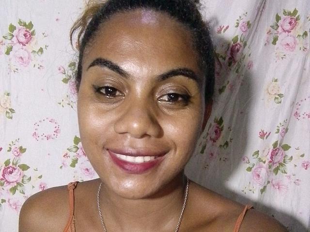 KennyReah has a shiny, brown-skinned face with thick eyebrows, brown eyes, and a pink smile. She's wearing an orange tank top and a silver necklace.