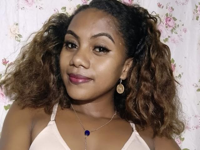 KennyReah has curly brown hair, medium brown skin, and wears a pink bra with a blue jewel necklace. She's smiling with pink lipstick, brown eyes, and gold earrings.