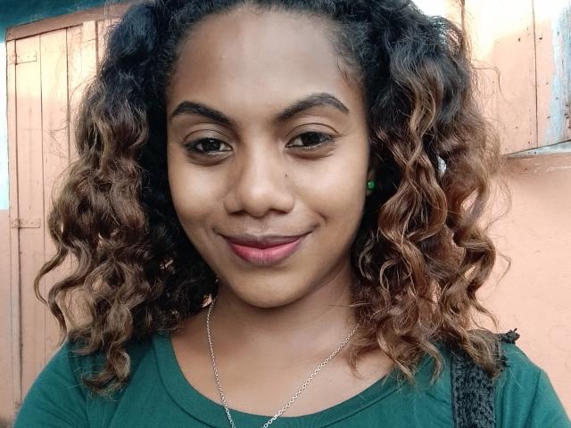 KennyReah has curly brown hair, medium skin, green eyes, and a small nose with a subtle smile. She's wearing a green shirt and a silver necklace.