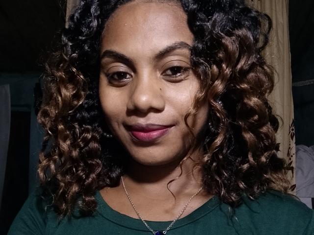 KennyReah has curly brown hair, dark brown eyes, and is smiling with pink lipstick. KennyReah is wearing a green shirt and a silver necklace. KennyReah's face is centered.