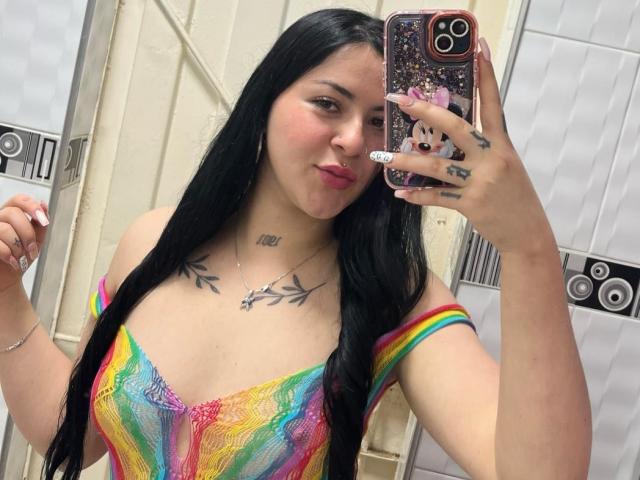 EmmieX with long black hair, pink lipstick, rainbow dress, showing off tattoos on chest and hand, taking a mirror selfie, holding a phone with a cute case.