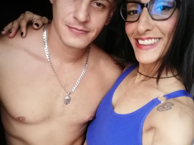 Shirtless SweetDangers with a silver necklace and nipple ring, standing next to a smiling SweetDangers in a blue tank top, showing a tattoo on their right shoulder.