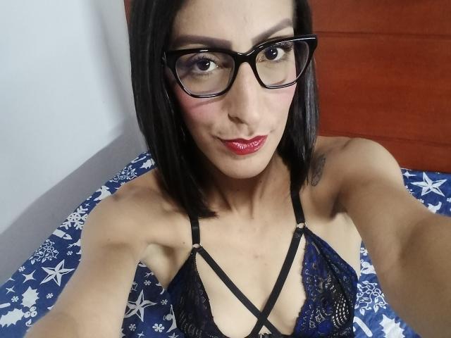 SweetDangers, wearing black lace lingerie with strappy details, has small tits, black glasses, red lipstick, and a cute smile, with short black hair and a tattoo on the arm.
