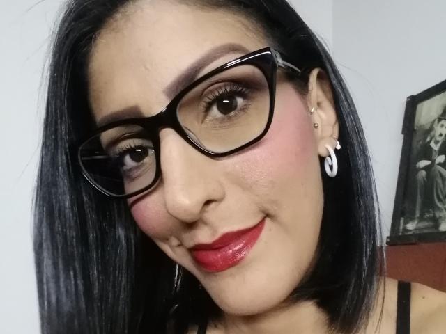 SweetDangers has dark hair, thick glasses, red lipstick, and a slight smile. She's wearing a black top with a small collar. Her nose is small, and she has earrings.