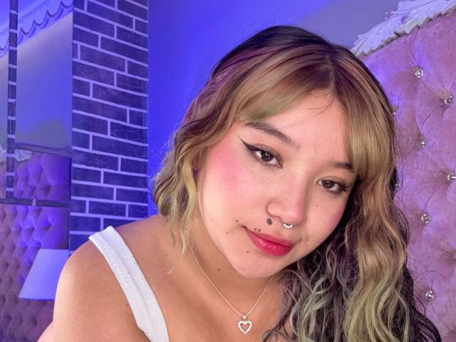 HonnyBlaice, met blond golvend haar, een neusring, een lipring en een hartvormige ketting, draagt een witte top, heeft roze blush en eyeliner, en een schattende glimlach. Slaapkamer met een roze gestoffeerd hoofdeinde..