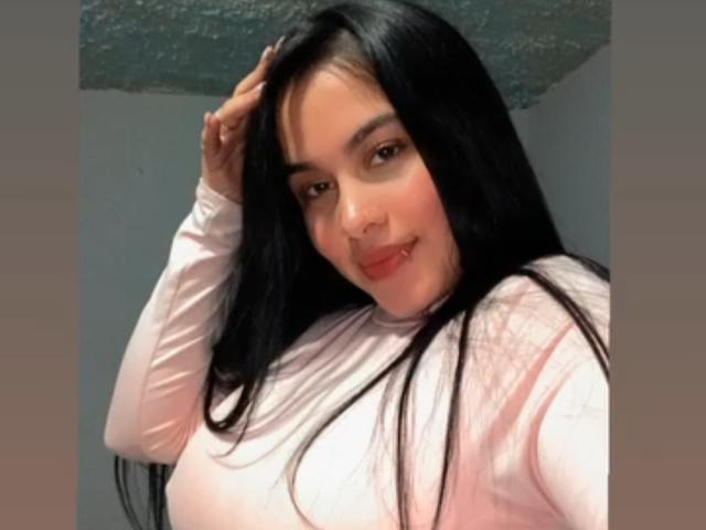 PerlaJones69 with long black hair, pink long-sleeve top, smiling, hand in hair, small red lips, cute, soft light, slightly flushed, medium breasts, casual selfie, relaxed.