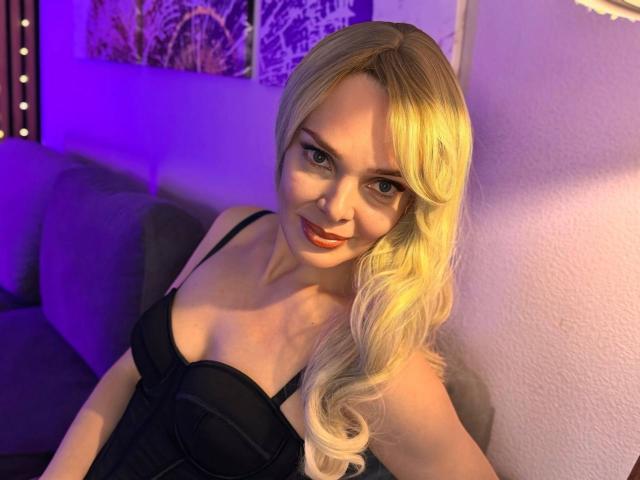 MoniDream has blonde hair, a cute smile, and is wearing a black corset that shows her small tits. She's sitting in purple-lit room, looking at the camera.