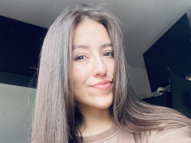 Katia Rogers tiene el cabello largo y lacio, de color marrón, piel clara y está sonriendo con labios rosados. Lleva una camisa marrón y tiene una pequeña peca debajo del ojo derecho..
