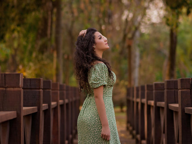 CaskiallX, with curly dark hair, green dress, and small tits, stands on a wooden bridge, hand in hair, looking up, surrounded by brown railings and green trees.