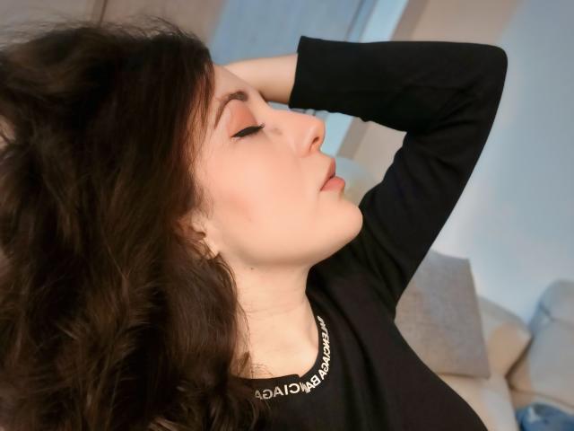 MabyLegrand with dark wavy hair, black long-sleeve shirt, eyes closed, arm raised, head tilted back, showing off pale skin and subtle makeup. Her lips are slightly parted.