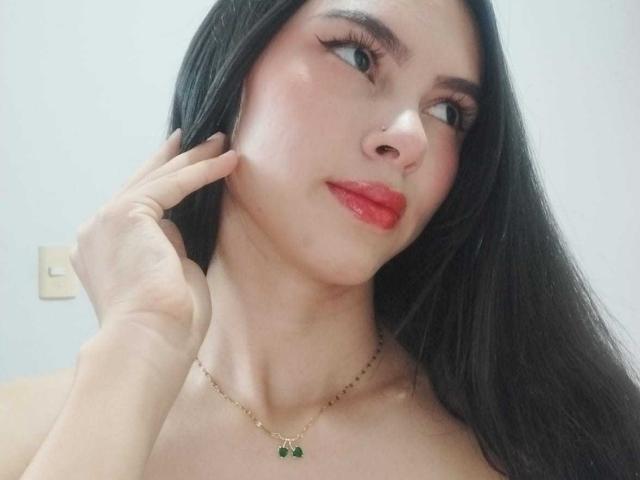 AngellAdamns hat langes, schwarzes Haar, roten Lippenstift und eine goldene Halskette mit grünen Steinen. Sie berührt ihre Wange mit einer Hand und blickt leicht lächelnd zur Seite..