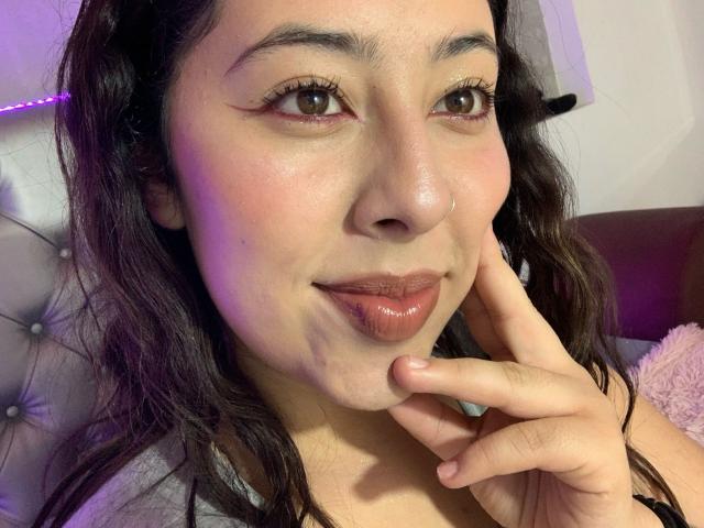 ScarlettParchMusa with wavy dark hair, brown eyes, and pink lips, touching her chin, showing a subtle smile, wearing a grey top, and a nose ring.