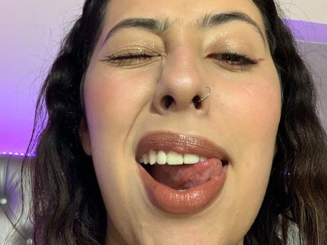 ScarlettParchMusa with eyes closed, tongue out, nose ring, and glossy pink lips, showing white teeth, wearing subtle makeup, and having wavy dark brown hair.