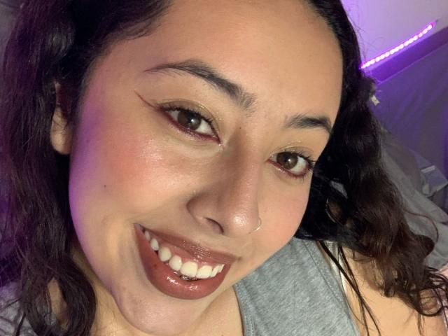 ScarlettParchMusa with dark wavy hair, brown eyes, and a cute smile showing glossy brown lips. She's wearing a gray shirt and has a small nose ring.