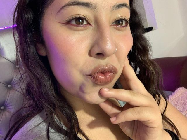 ScarlettParchMusa with puffy pinkish lips, dark wavy hair, brown eyes, and a finger touching her chin, looking slightly to the side, showing subtle pink blush on her cheeks.