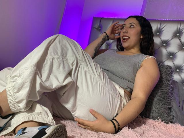 ScarlettParchMusa, curvy with a big ass, wearing a gray tank top and white pants, lying on a pink fuzzy bed, smiling, hand on face, tattoos visible, purple light.