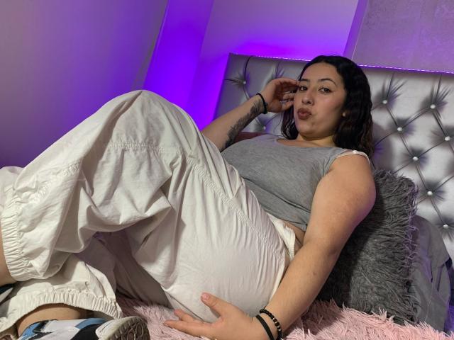 ScarlettParchMusa with puffy white pants, gray sleeveless top, and tattoos, laying on a bed, pouting, wearing a black bracelet, with a fluffy pink blanket.